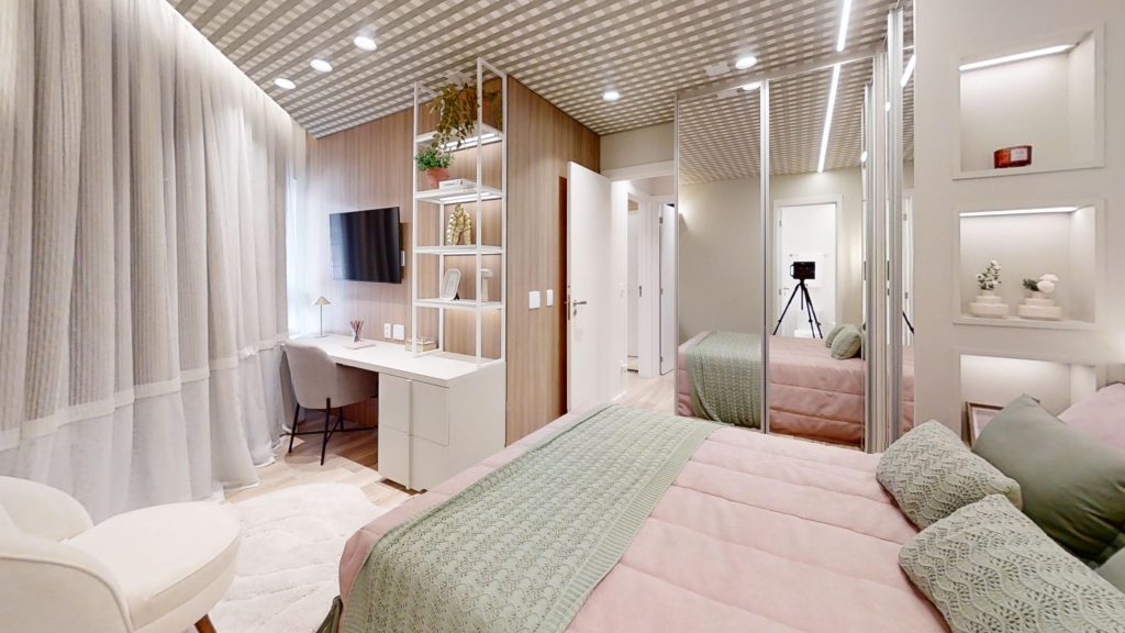 Um qurto decorado no empreendimento de alto padrão AYA, em Guarulhos, com cama de casal, espelhos, penteadeira, televisão poltrona e cortinas 